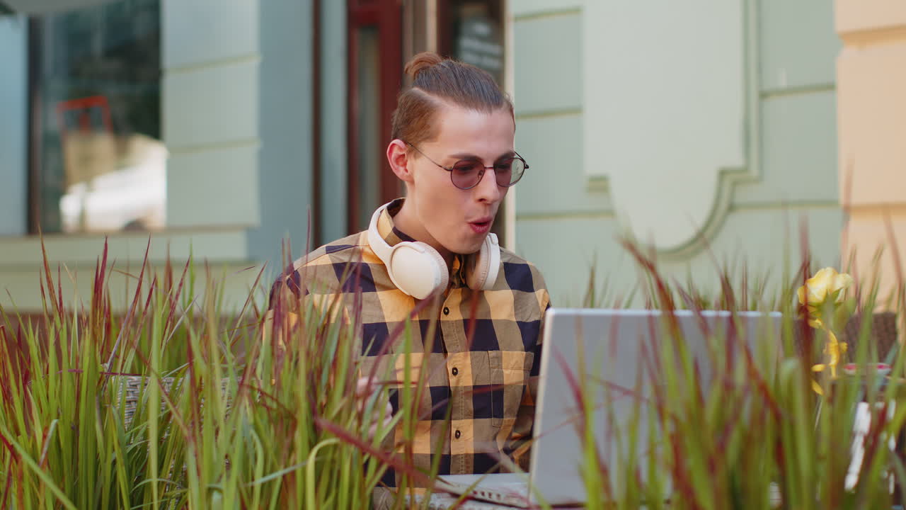 Glad mandlig turist bruger bærbar computer og fejrer gode nyheder på en caféterrasse i byen.