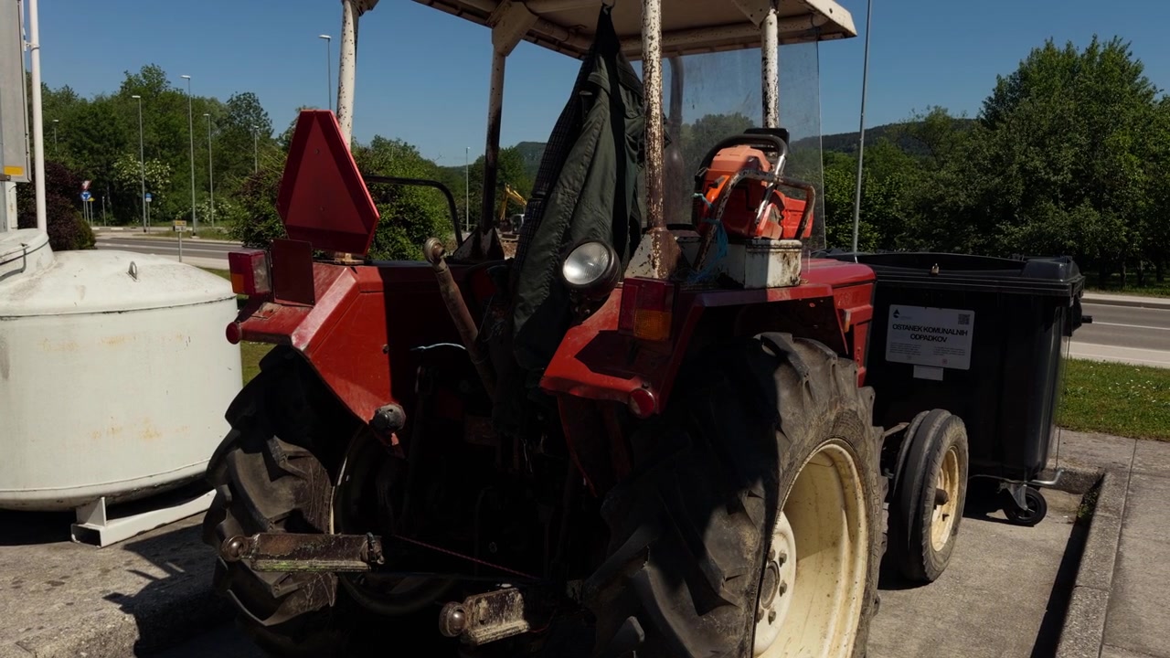 Gammel rød traktor parkeret ved siden af genbrugscontainere på en solskinsdag i Europa. Landbrugskøretøj brugt til landbrug, vedligeholdelse og landbrugsarbejde udendørs.