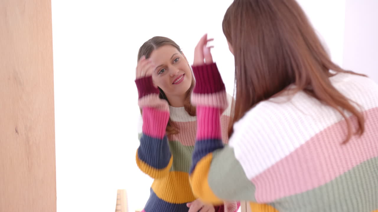 Smiling young woman in colorful sweater looking at herself in mirror