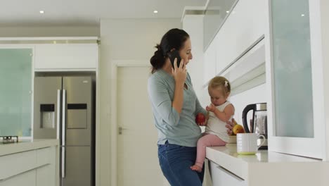 caucasian mother holding her baby talking on smartphone in the kitchen at home