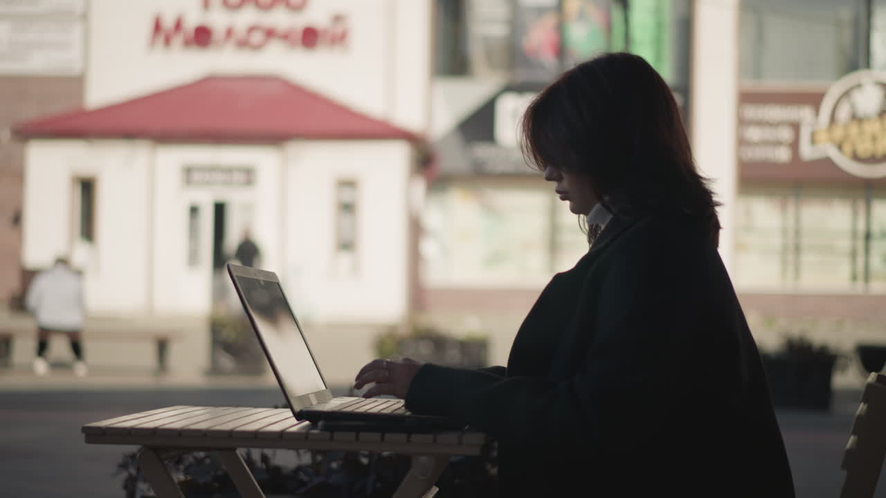 Woman in black coat working on laptop at outdoor table in urban environment, with blurred background of red-roofed building and passerby in motion