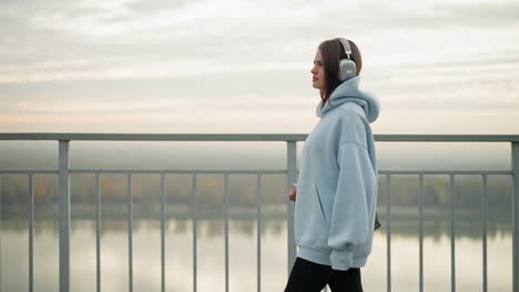 side view of female adult walking slowly while listening to music with headphones, enjoying peaceful outdoor walk in urban setting, calm atmosphere, serene surroundings by river and rail