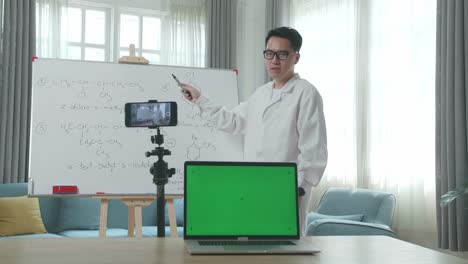 asian male teacher with glasses and green screen laptop shoots video by smartphone camera while teaching chemist at home