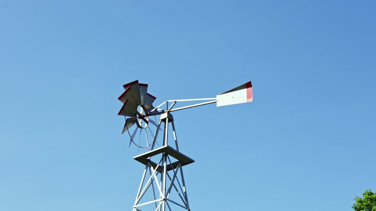 Un mulino a vento si erge imponente contro un cielo azzurro limpido durante una giornata di sole in una zona rurale, mostrando le sue pale pronte a catturare il vento e produrre energia