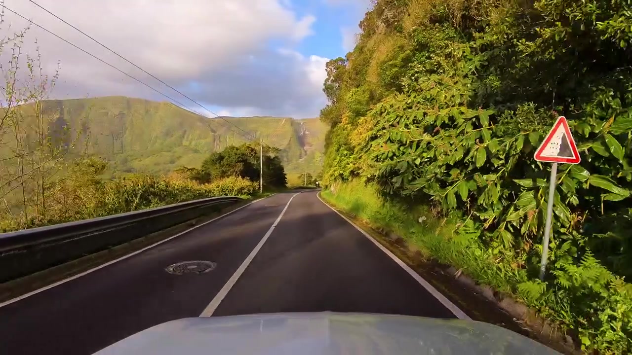 Percorso panoramico in auto lungo una strada di montagna con cascate al tramonto e rigogliose scogliere vulcaniche vicino a Poco da Ribeira do Ferreiro nelle Azzorre, Portogallo.