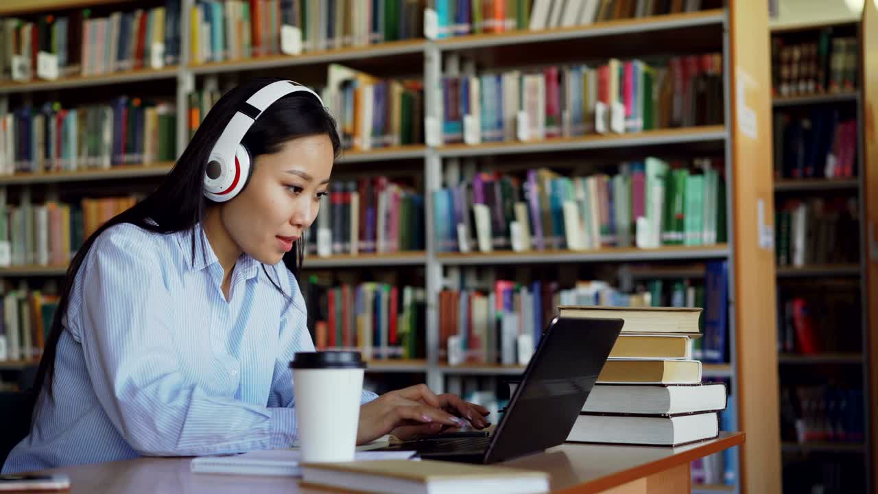Beautiful asian female student listening music in headphones sitting at desk in library working at school project. Young girl typing on laptop, drinking coffee and writing in copybook