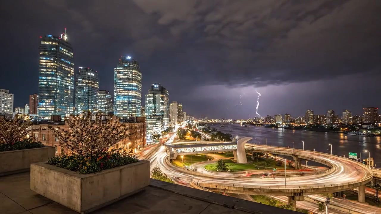 A stunning visual contrast of city life at sunset and thunderstorm, showcasing skyscrapers, river, and illuminated roads beneath a dramatic sky.
