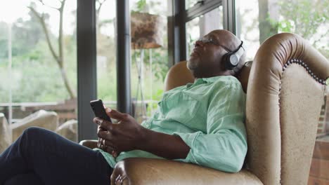 happy african american senior man relaxing in armchair, listening using headphones and smartphone