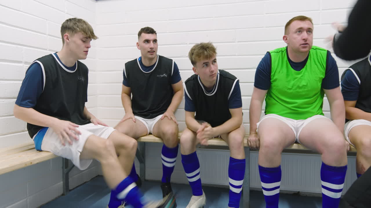 Male soccer players wearing black vests and tying shoes sitting on bench in changing room with coach