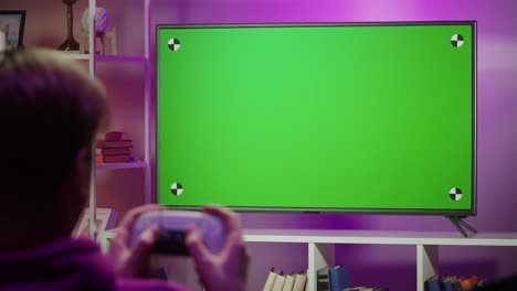 man playing video game in living room in evening. young guy sitting on sofa against tv with chroma green screen. student gaming with modern console.