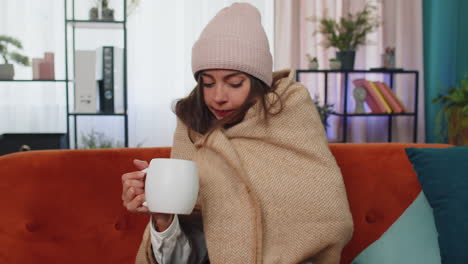 sick young woman wear hat wrapped in plaid sit alone shivering from cold on sofa drinking hot tea