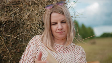 primer plano de una mujer con una camiseta de patrón de red con gafas de sol en la cabeza, profundamente enfocada en leer un libro, se apoya contra una bala de heno en un campo abierto con árboles en un fondo suave