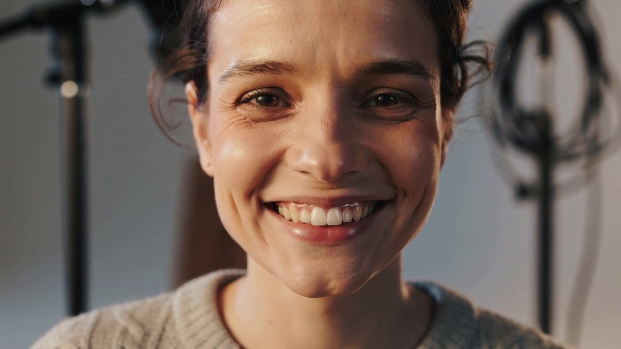 Smiling knitwear woman holding gaze as camera starting recording in studio, mic stand, cable behind