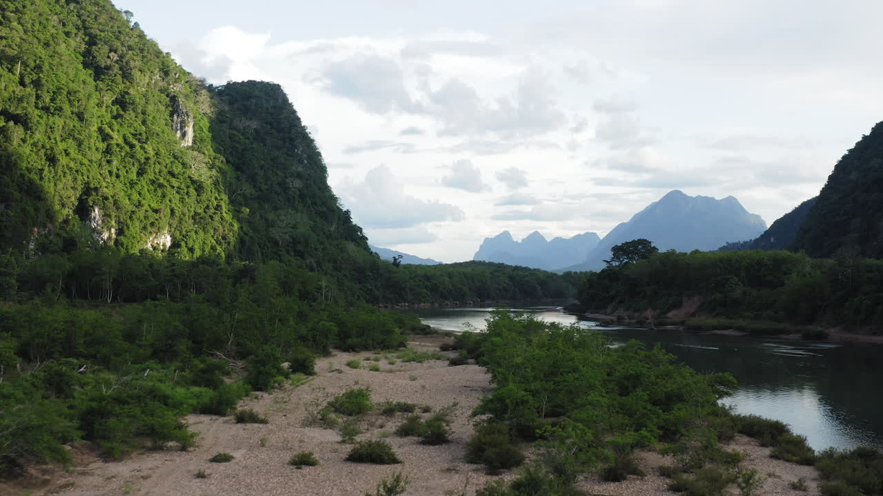 Drone che vola a bassa quota avanzando da un terreno sabbioso, inclinandosi verso il fiume e la montagna illuminata dal sole