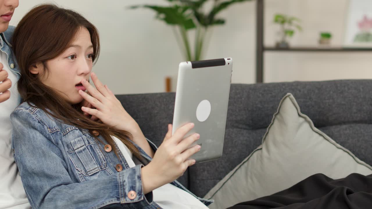 明るいリビングで、恐ろしいタブレット動画を見て驚いている若いアジア人カップル