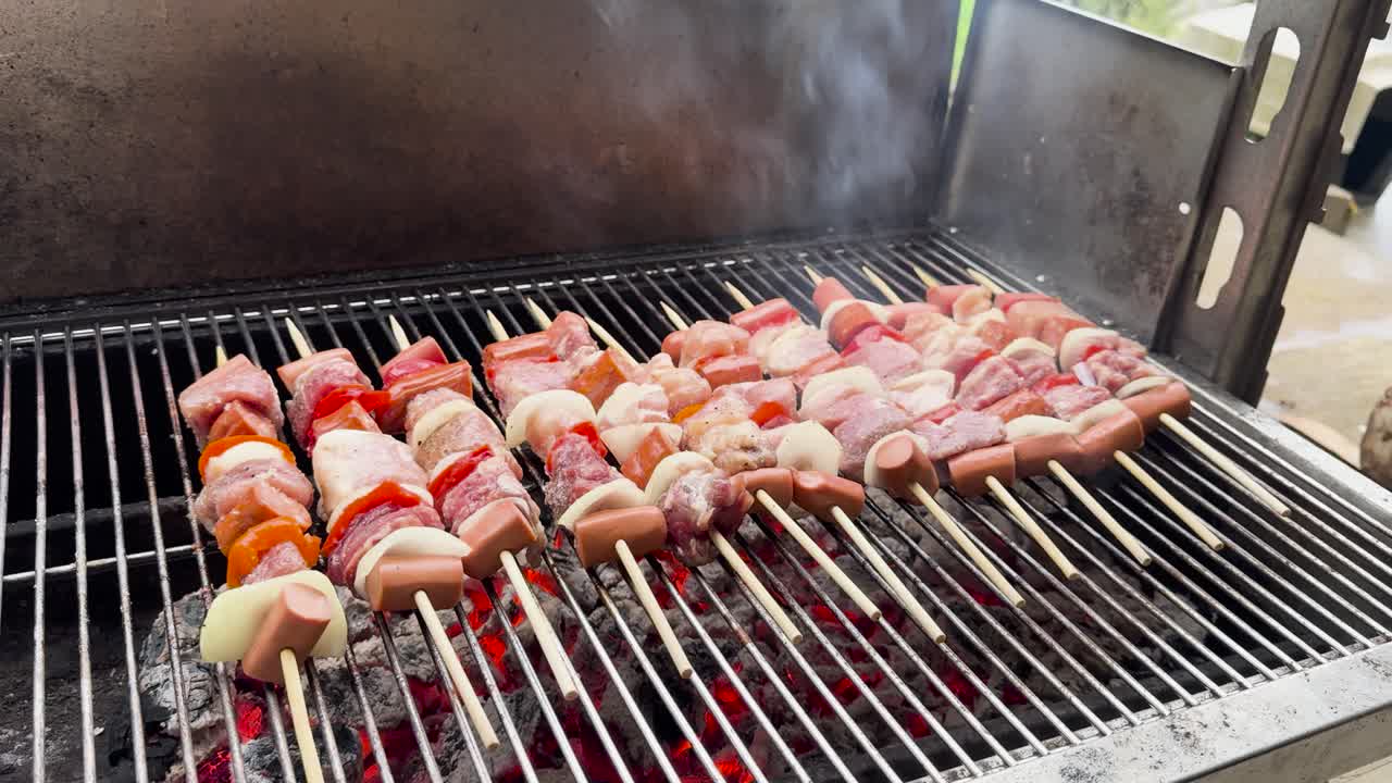 Handheld video of meat skewers grilling on a charcoal barbecue, with red peppers, white onions, and sausages. Glowing red coals and rising smoke create a rustic, savory scene.