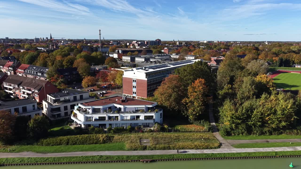 Seitliche Luftaufnahme von deutscher Stadtarchitektur im Herbst mit rot gedeckten Häusern, modernen Gebäuden und bunten Herbstbäumen unter einem klaren, blauen Himmel. Weitwinkelaufnahme. Grünes Flusswasser im Vordergrund