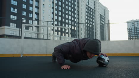 young man performing dynamic push-ups alternating hand placement on soccer ball, showcasing strength, coordination, and agility, background includes goal post and residential buildings