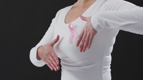 Mid-section-of-a-woman-showing-the-pink-ribbon-on-her-chest-against-black-background