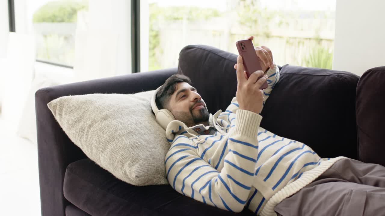 Uomo indiano rilassato su un divano con le cuffie che usa lo smartphone per scorrere il feed.