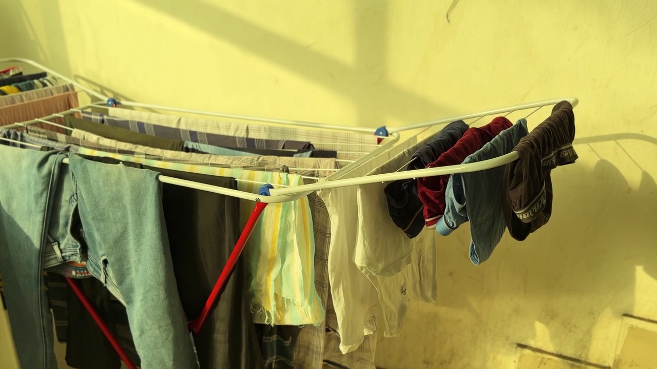 Clean laundry hanging on a white drying rack outdoors on a sunny day, with warm daylight casting shadows against a textured yellow wall. Perfect for daily routines and household chores concepts.
