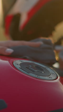 un primer plano de una mano limpiando suavemente el tanque de combustible rojo de una motocicleta usando un paño