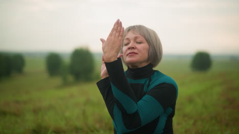 mujer de mediana edad en traje verde y negro practicando yoga al aire libre en postura de cara de vaca, con los ojos cerrados, centrándose en la atención y la respiración en un campo de hierba sereno bajo un cielo nublado