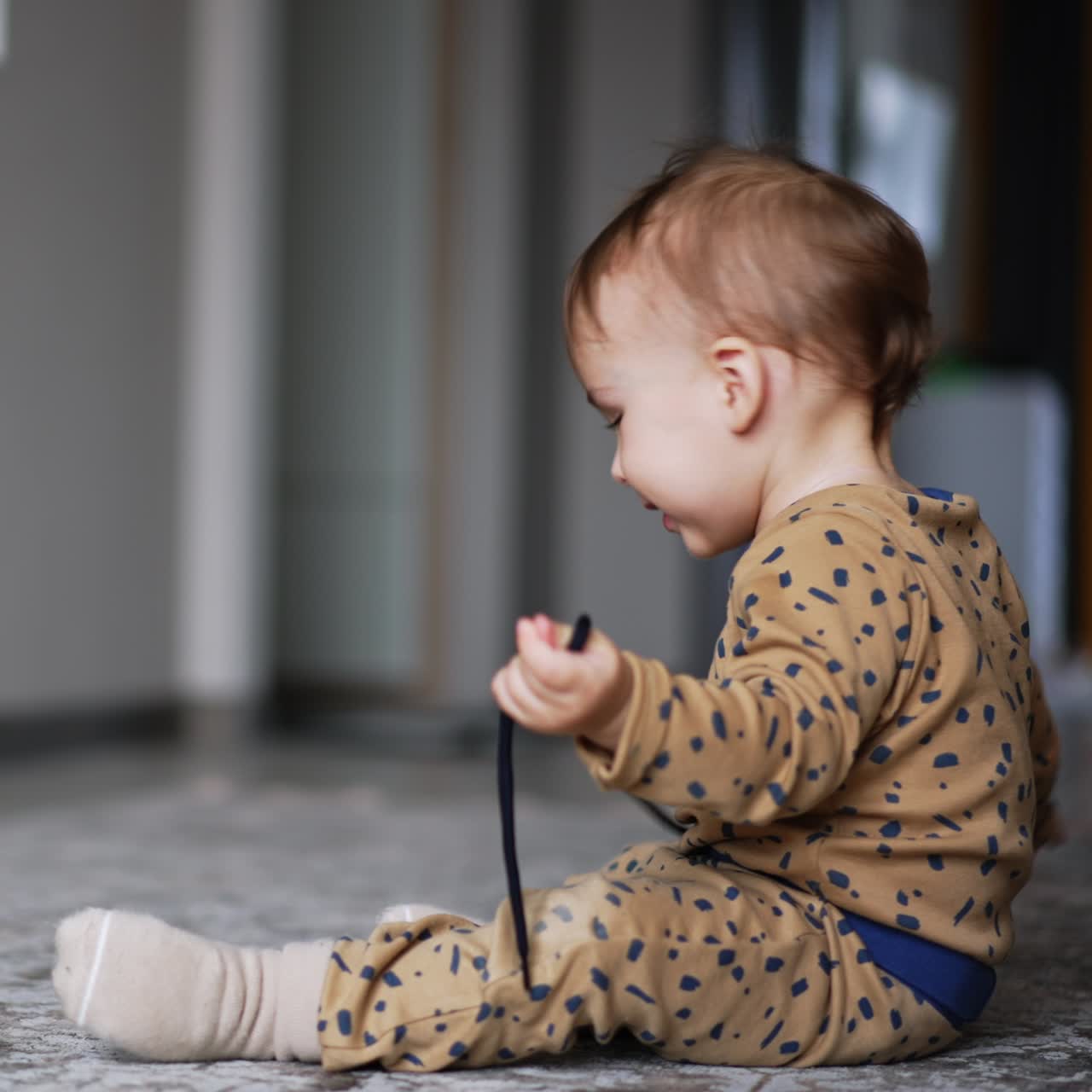 Cute one year old boy sits on the floor playing with a cord. Funny kid looks into camera and crawls away