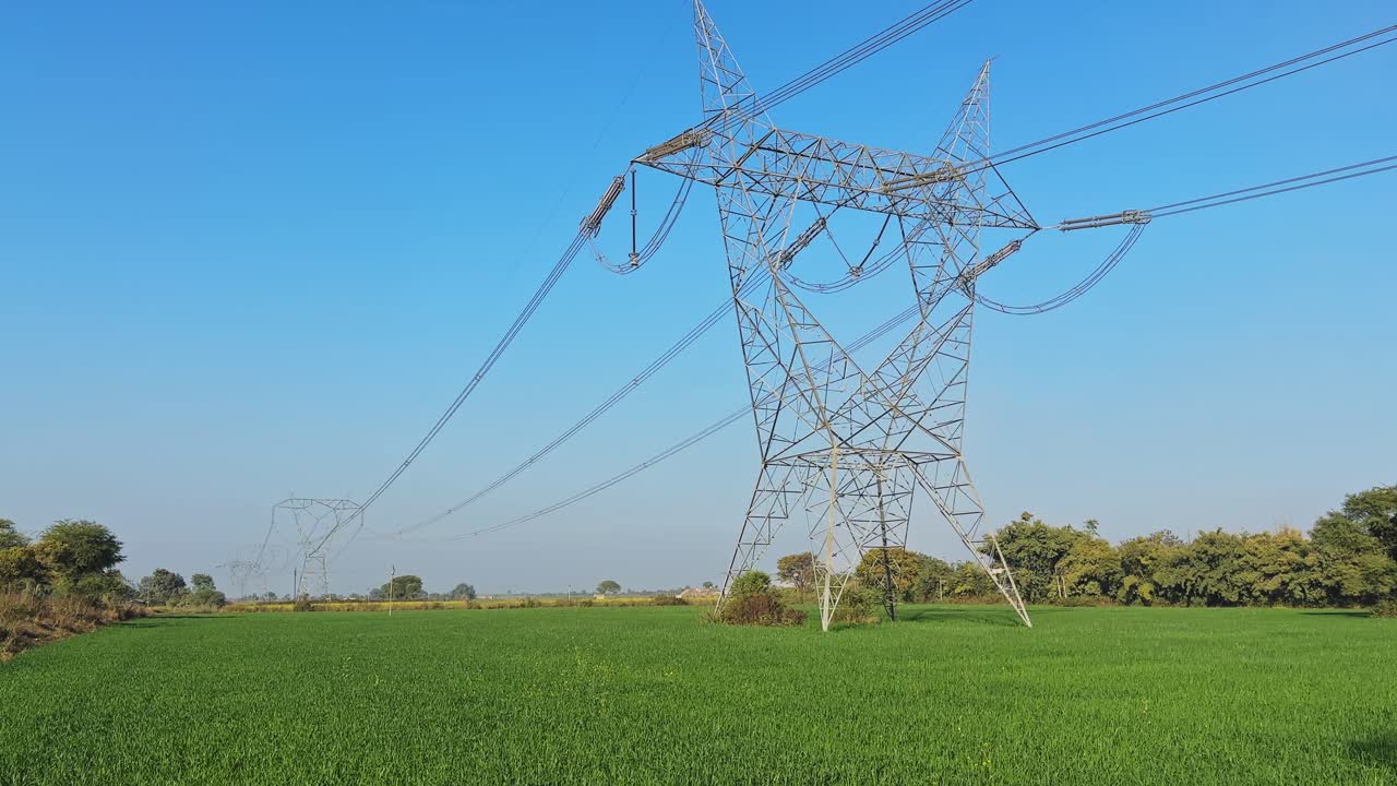 Tilt up shot starting from a lush green wheat field and revealing a high voltage transmission tower, power lines stretching across open farmland under a clear blue sky