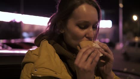 Hungry-young-woman-with-sparkles-on-her-face-is-taking-a-hamburger-outside.-Pleased.-Side-view