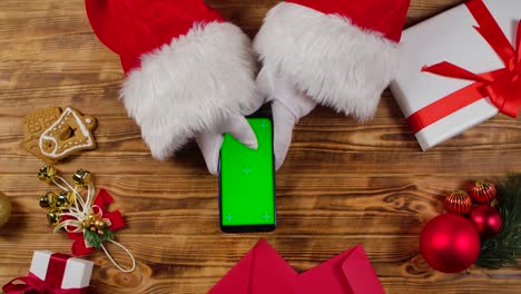 santa claus usa un teléfono inteligente con pantalla verde llave de croma por madera mesa decorada para el año nuevo. santa claus tocando en la pantalla para ver contenido, hacer compras en línea. cerrar. cámara lenta