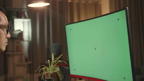 asian male works with mock up green screen computer display. he works at night
