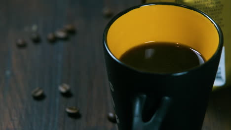 close up shot of a cup of coffee next to a packet of coffee beans and coffe beans on a wooden table