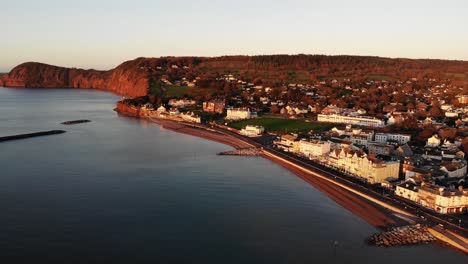 Toma-Aérea-De-La-Ciudad-De-Sidmouth-Y-La-Costa-Jurásica-Del-Suroeste-De-Inglaterra-Que-Refleja-El-Color-Rojo-Dorado-Del-Hermoso-Amanecer-De-La-Mañana