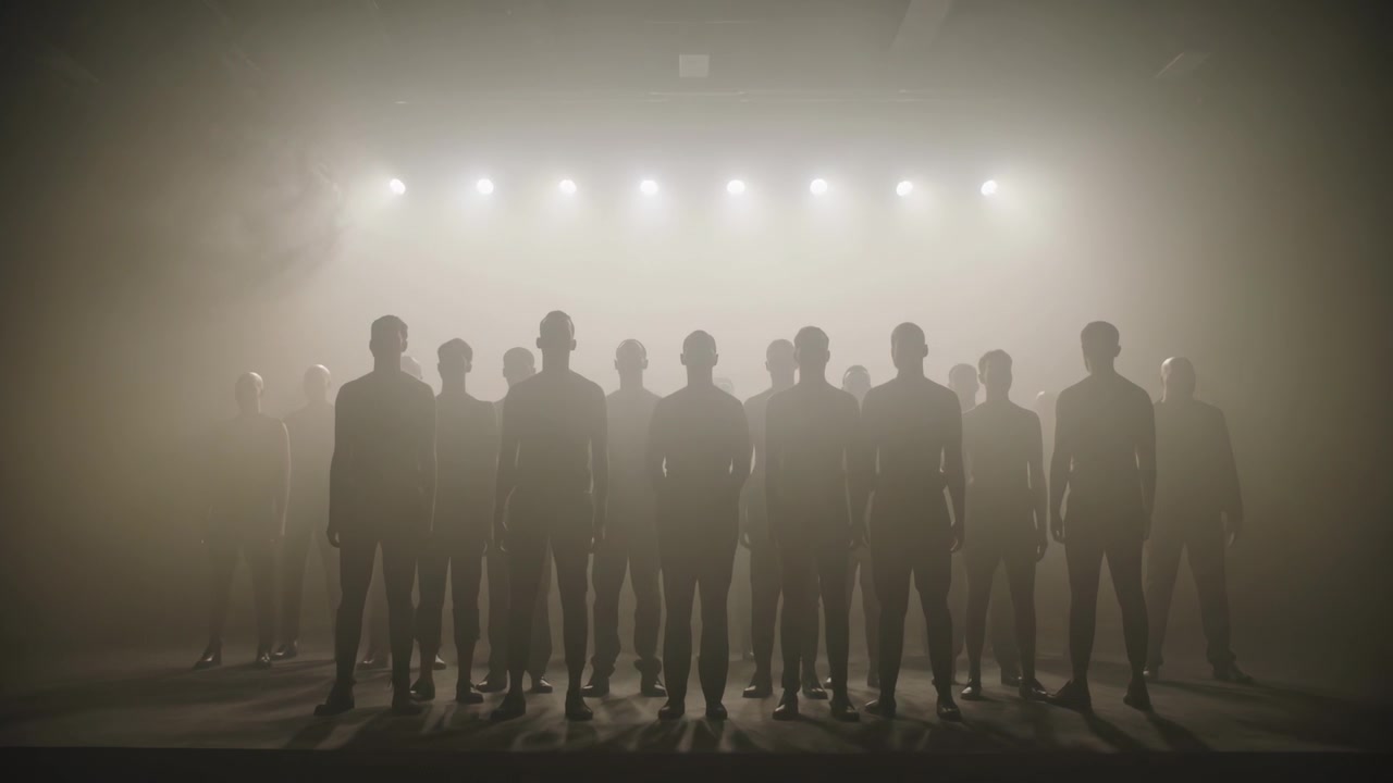 Gruppo in uniforme in piedi che mantiene la posizione su un palco poco illuminato, con luci a fila che si accendono tra la foschia per creare drammaticità.