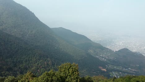 Flying-over-the-edge-of-a-mountain-to-reveal-the-valley-and-city-below-before-turning-to-the-left-to-see-the-mountains-on-a-hazy-day