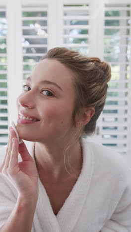 vertical video of biracial woman cleansing face with cotton pad in bathroom, slow motion