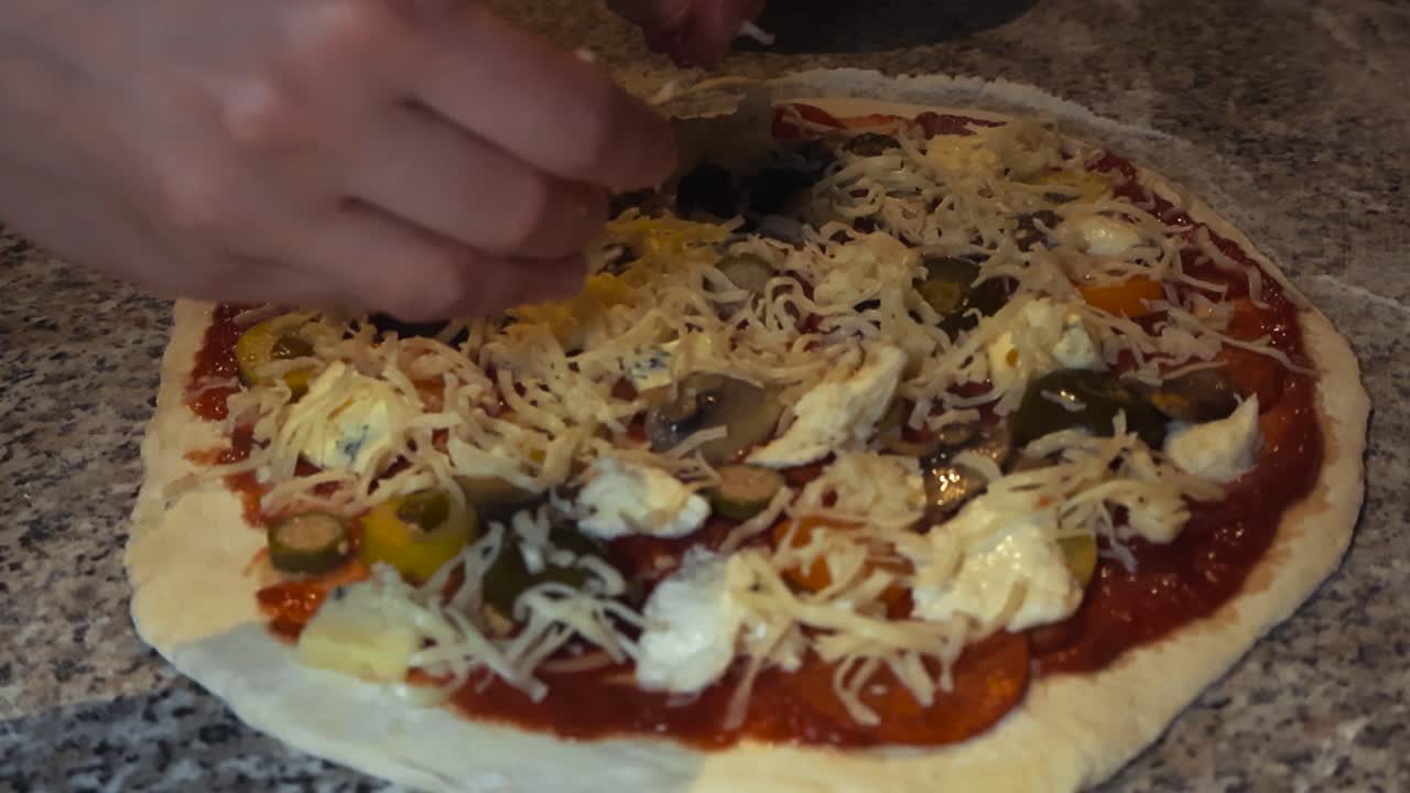 Slow motion video of a chef or a home cook spreading cheese or mozarella on top of freshly made pizza dough where meat, jalapenos and tomato sauce is visible. White flour around on the kitchen table