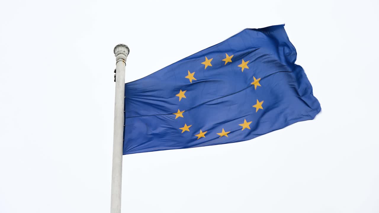 Slow-motion footage of the European Union (EU) flag fluttering atop a tall post, swaying in the wind on a cloudy day, capturing the flag's elegant movement as it reacts to the breeze.