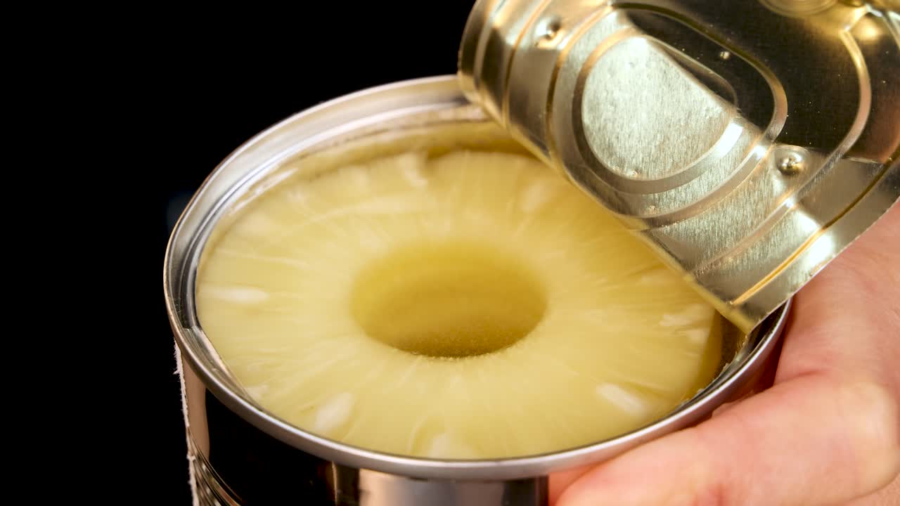 A hand opens a metal can of pineapple rings, revealing fruit in syrup. The scene is brightly lit with a close-up perspective and steady camera