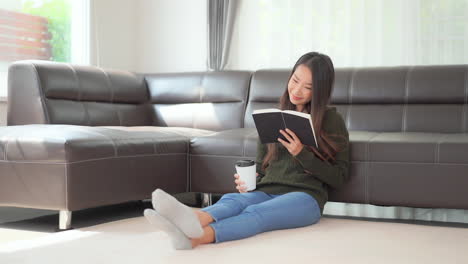 a pretty young woman sitting on the floor holding a cup of coffee while she reads a book