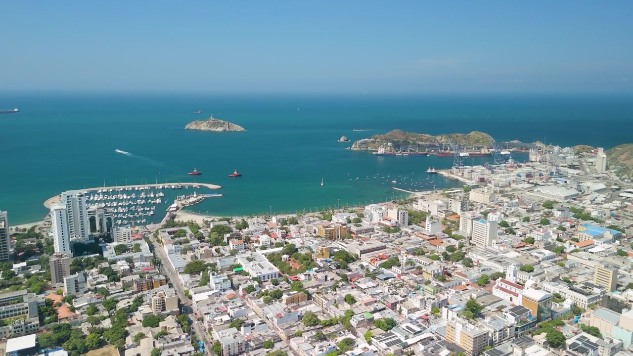 Sobrevoo aéreo de alta altitude sobre a cidade de Santa Marta revelando marina, Baía de Santa Marta, ilha do farol de Isla El Morro e porto industrial