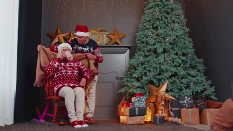 una pareja de ancianos celebra la navidad. el abuelo cubre con una manta a la abuela. la mujer bebe té.