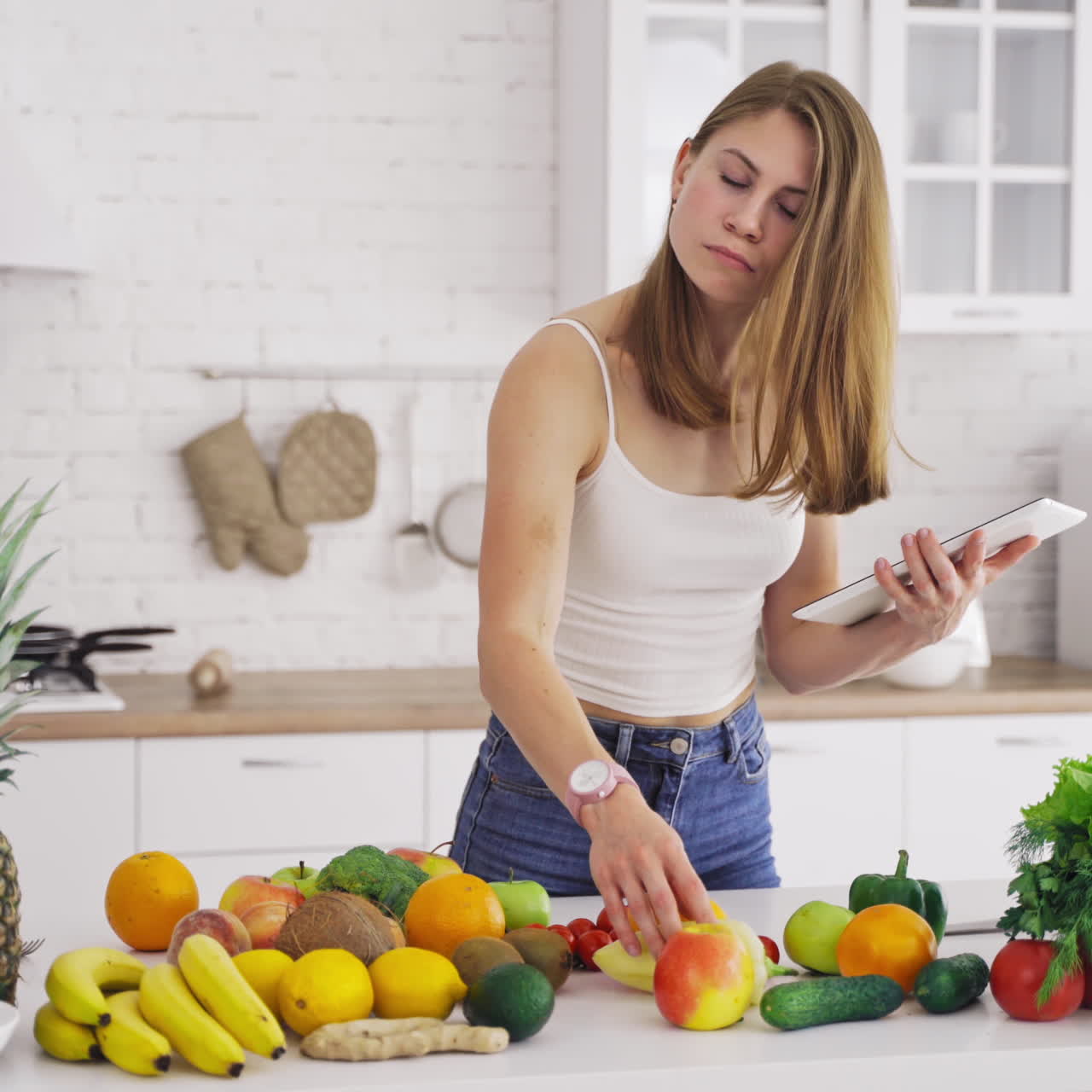 Beautiful woman looking into tablet on the light kitchen background. Young housewife chooses necessary organic fruit and vegetables for recipe to lose weight.