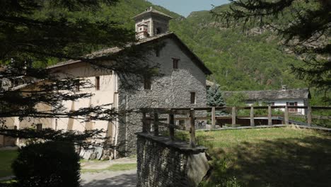 an old stone building with a bell tower is situated amidst lush green trees, set against the backdrop of a mountain range under a clear, sunny sky