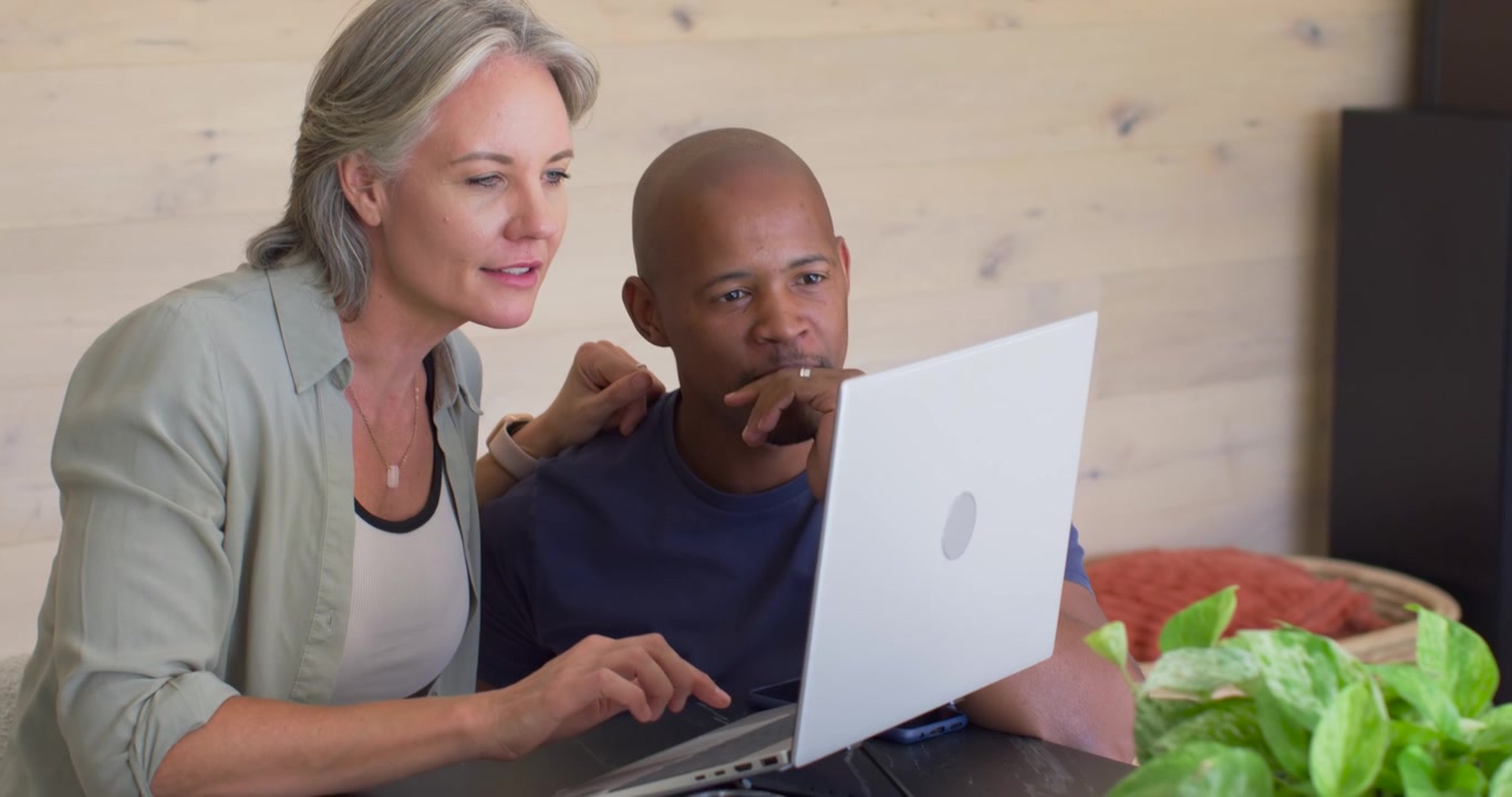 Diverse couple leaning after woman reaching, typing on laptop at home, man reviewing documents