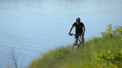 cyclist looks away. man standing with bike on a rock near river and enjoying landscape