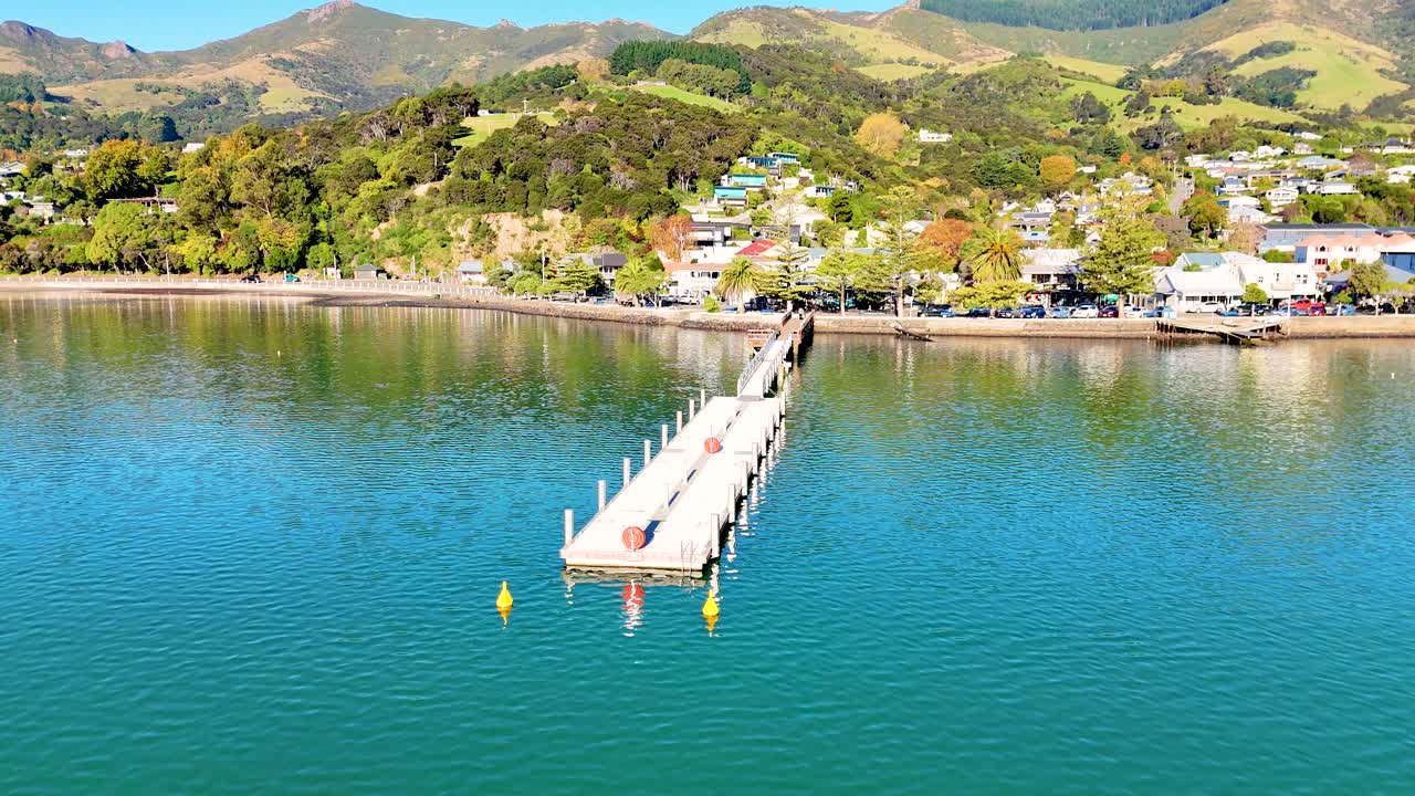 Drone footage captures Akaroa's scenic pier and harbor under clear skies, highlighting vibrant landscapes and tranquil waters
