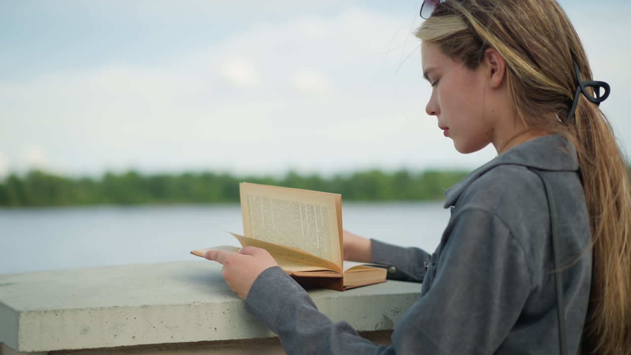 Lady wearing a grey shirt reading a book while resting on a fence, the wind gently lifts the pages of the book as she focuses on reading, with a background of greenery