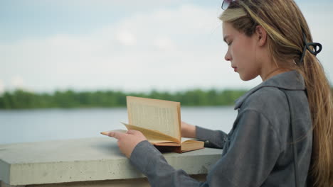 lady wearing a grey shirt reading a book while resting on a fence, the wind gently lifts the pages of the book as she focuses on reading, with a background of greenery